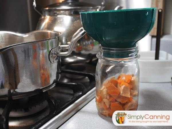 A canning jar filled half of the way with solid chicken and vegetable ingredients with a pot sitting to the side on the stovetop.