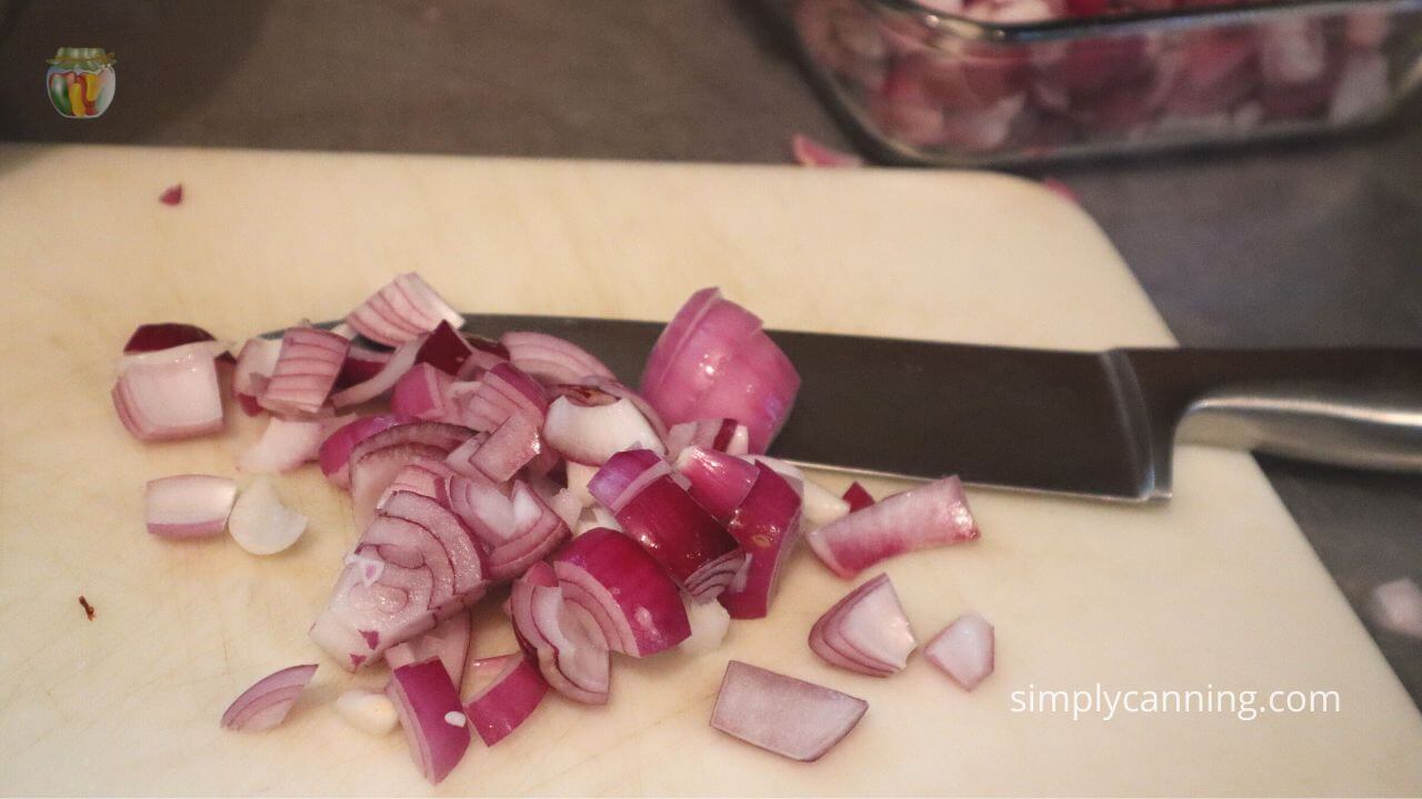White cutting board with chopped red onions and a large knife, clear baking dish filled with chopped red onions in the background