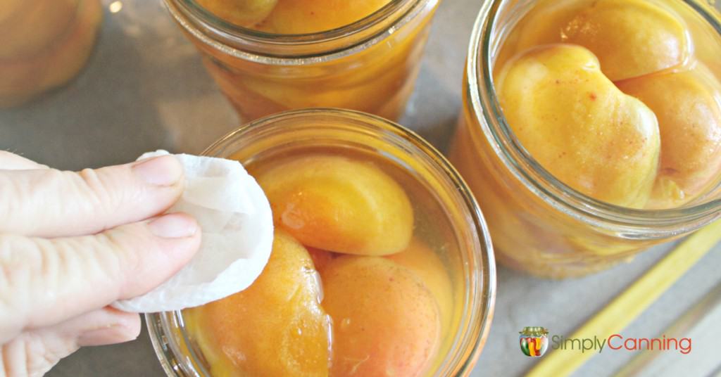 Wiping the rim of a jar filled with fresh apricot halves and syrup.