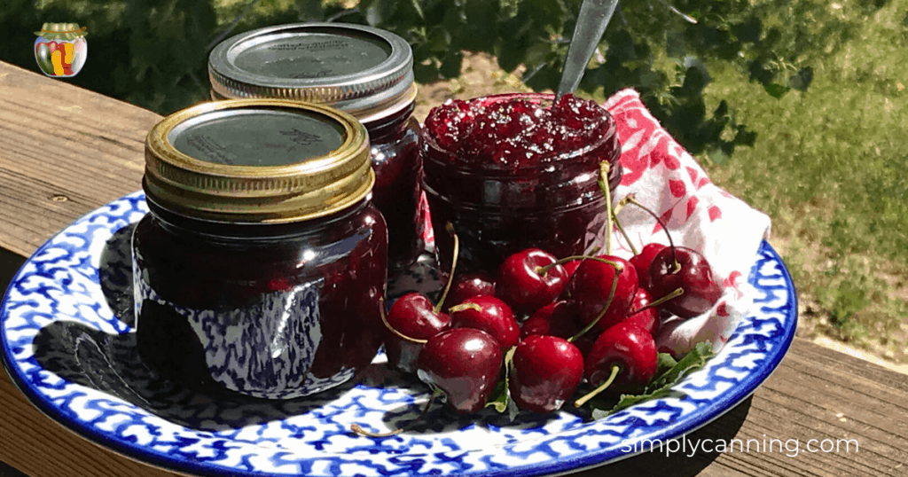 An open jar of cherry jam sitting on a plate surrounded by jars of more jam as well as fresh cherries.