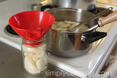 Packing slices of fruit into ready canning jars using a red canning funnel.