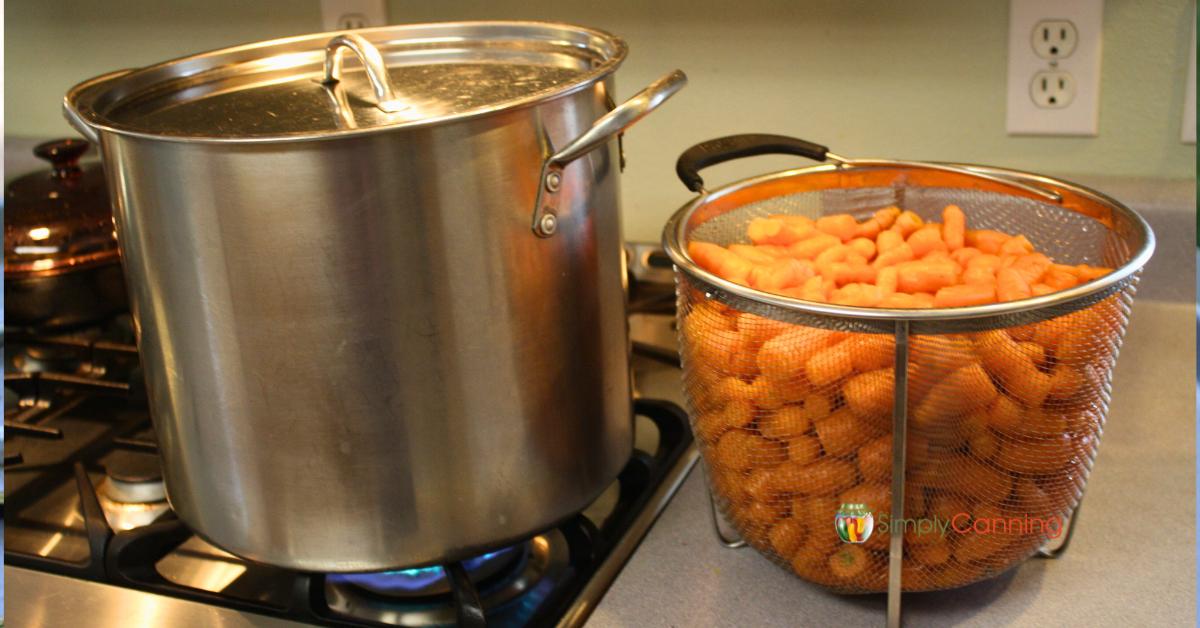 Aluminum stock pot being heated on a gas stove, a full basket of small carrots waiting to be blanched before freezing.