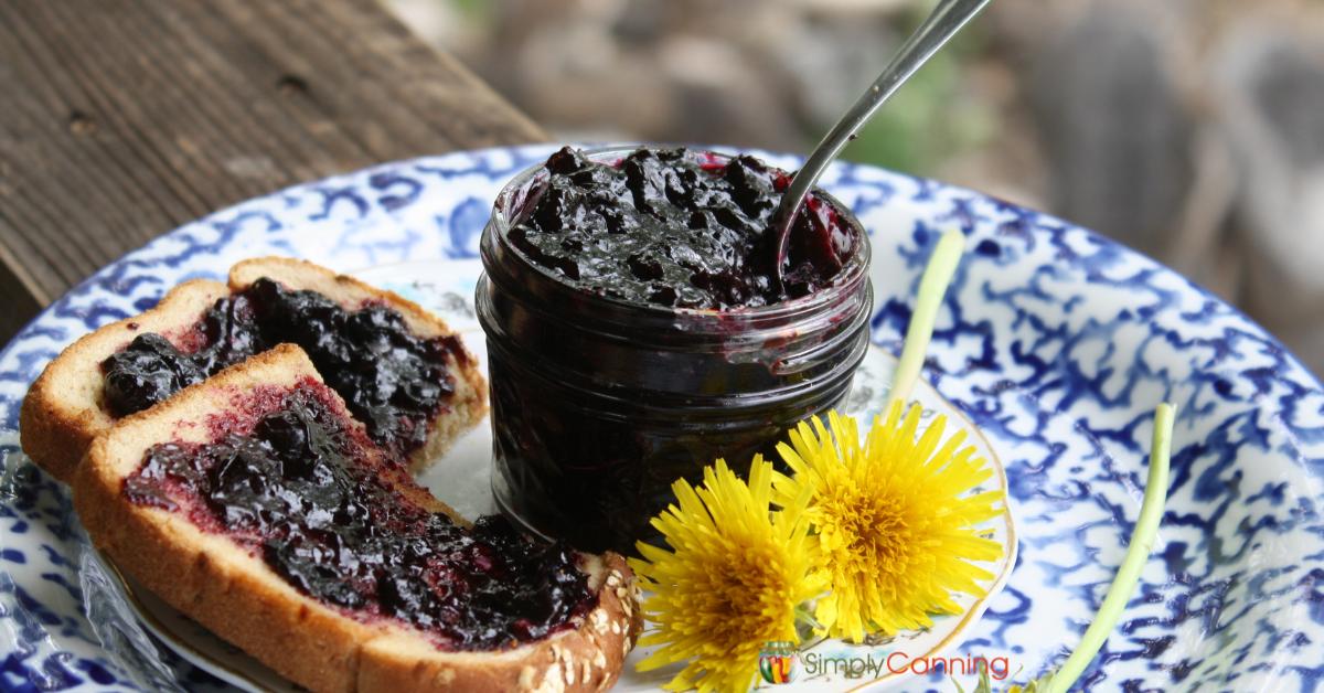 A small jar of thick blueberry jam with toast on the side that’s also spread with jam, dandelions decorate the pretty plate.