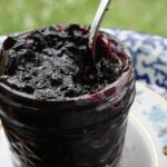 A full open canning jar of deeply colored blueberry jam sitting on a china plate.