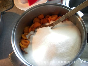 Pouring lots of white sugar into a pot of sliced apricots.