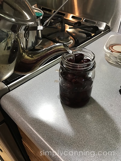 Pouring liquid from a tea kettle into jars of cherries.
