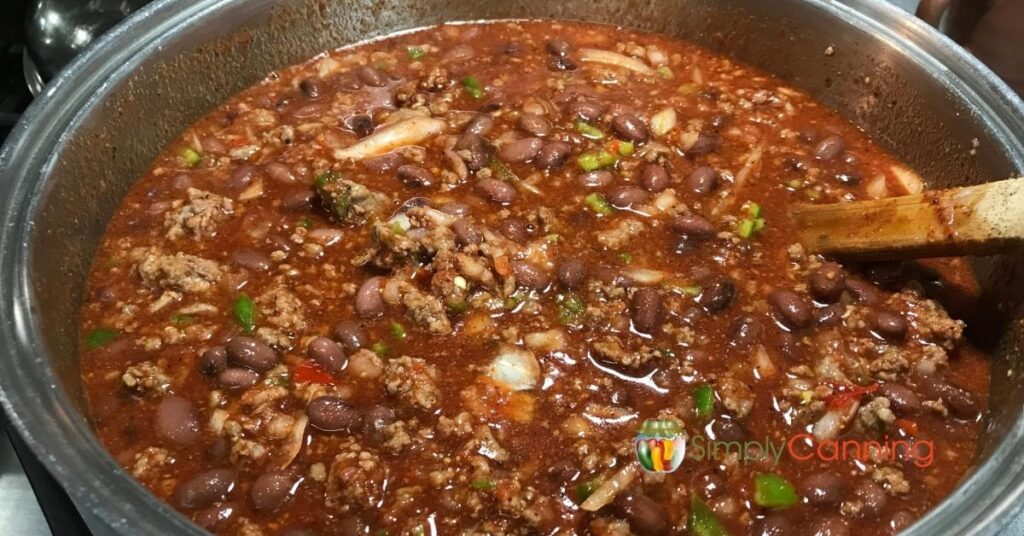 Top down view of a pot of chili with lots of ground meat, beans, chopped green pepper and onion, wooden spoon giving it a stir.