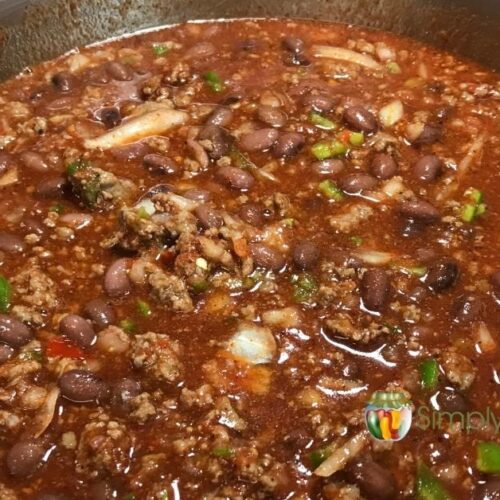 Top down view of a pot of chili with lots of ground meat, beans, chopped green pepper and onion, wooden spoon giving it a stir.