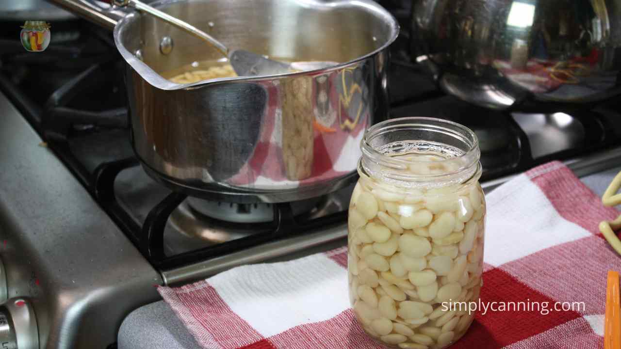Jar of lima beans on a red checked cloth, being filled with beans from the pot on the stove in the background. 