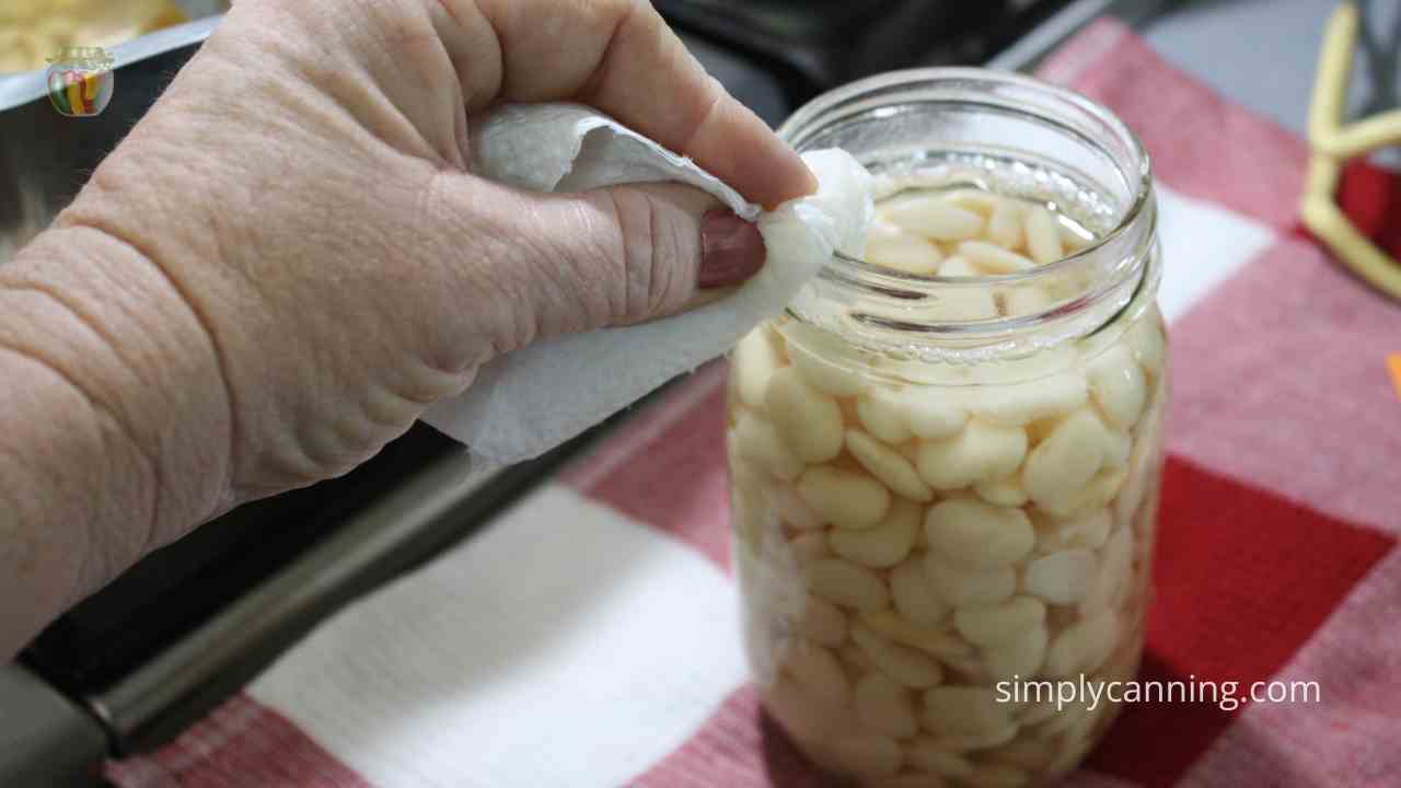 Jar of lima beans on a red checked cloth, a hand wiping the rim with a paper towel.  