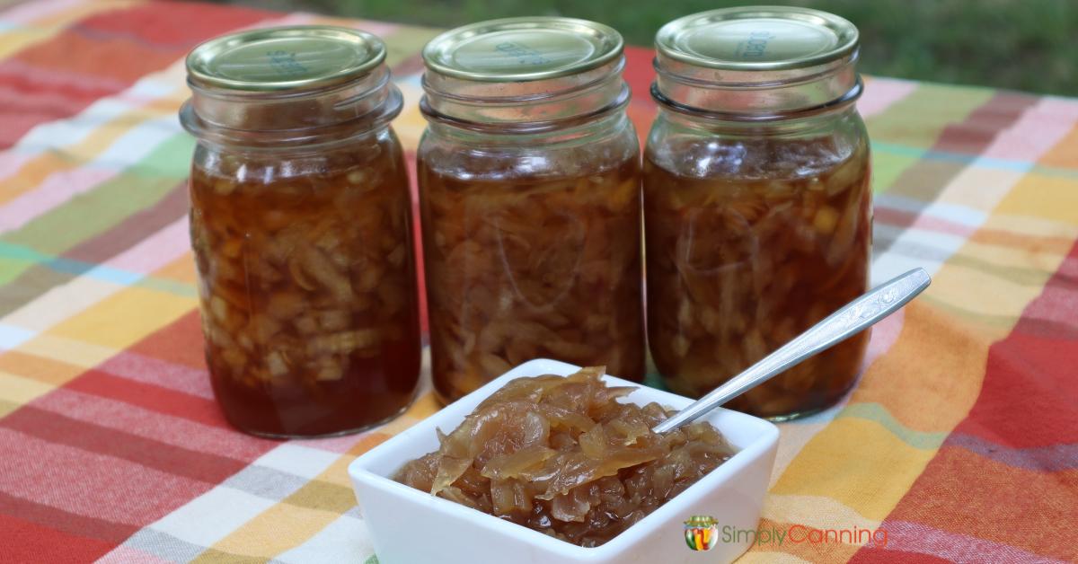 Autumn colored table cloth with white dish of onions and honey sauce, three pint jars of canned onions in the background.