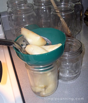 Packing pear slices into clean canning jars through a green canning funnel with other canning jars in the background.