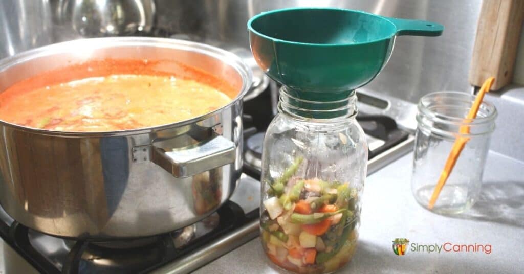 Canning jar filled with solid soup ingredients with a boiling pot of soup to the side.