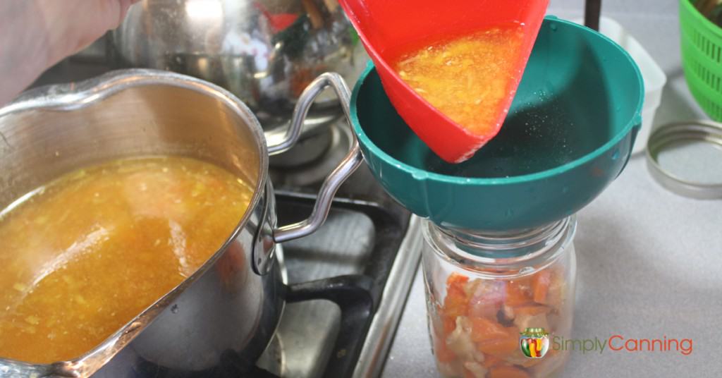 Pouring soup into a canning jar through a canning funnel with the pot of soup to the side.