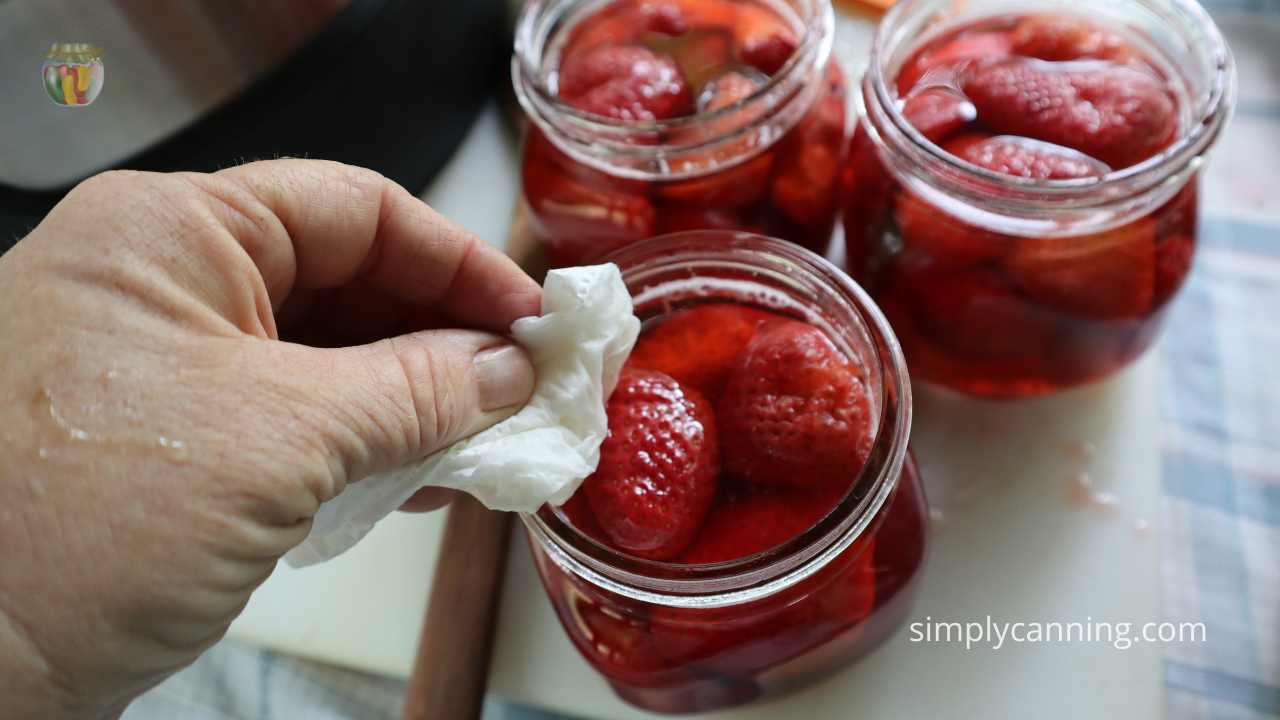 3 pint jars of strawberries on a white cutting board with the rims being wiped clean with a paper towel. 