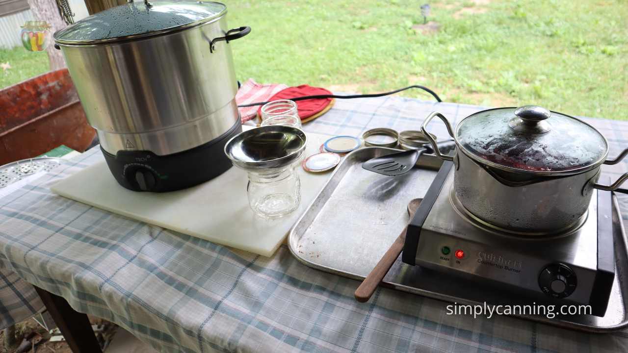 Table top with a light blue plaid tablecloth, electric canner, countertop burner and other hand canning tools lying about.