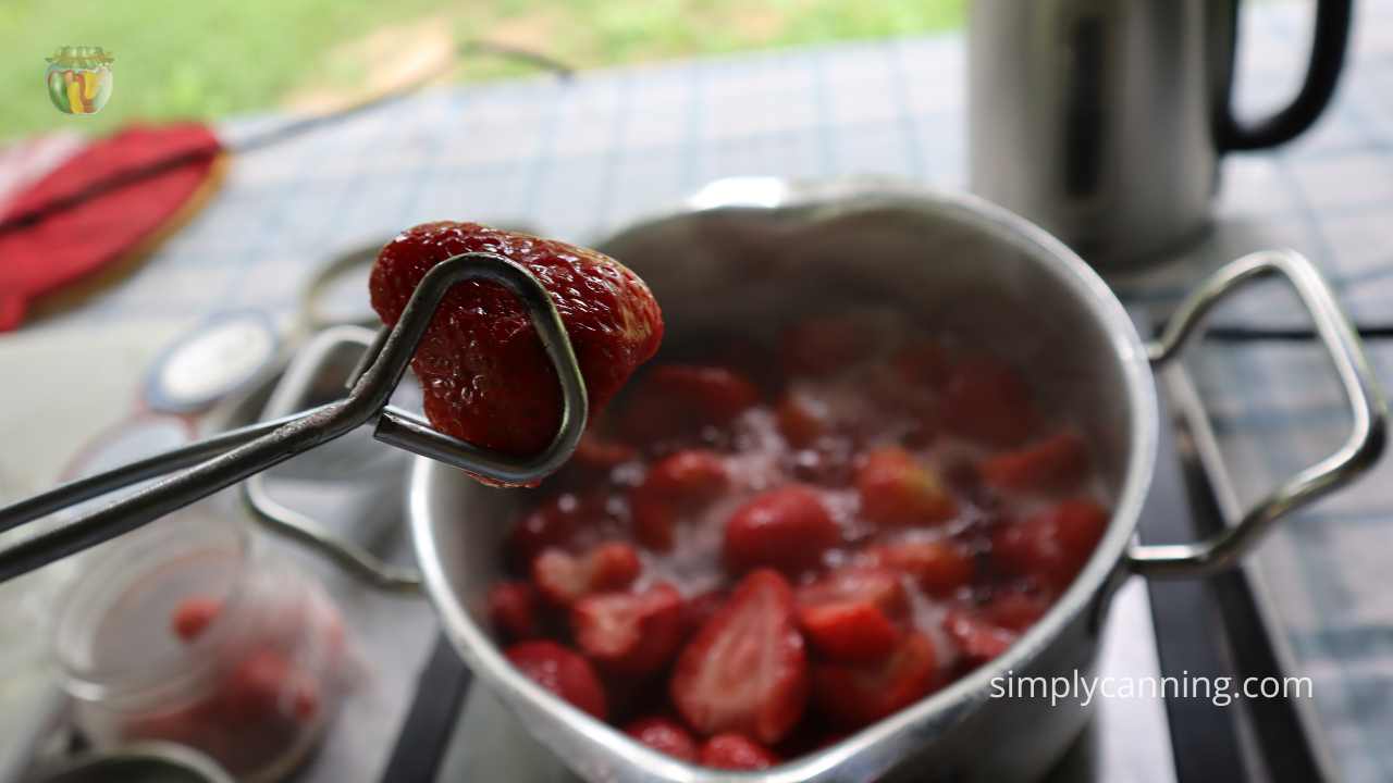 Close up of tongs holding a strawberry above a pot of cooking berries.  