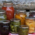 Jars of colorful home canned food sitting on a gingham cloth in front of a log background.