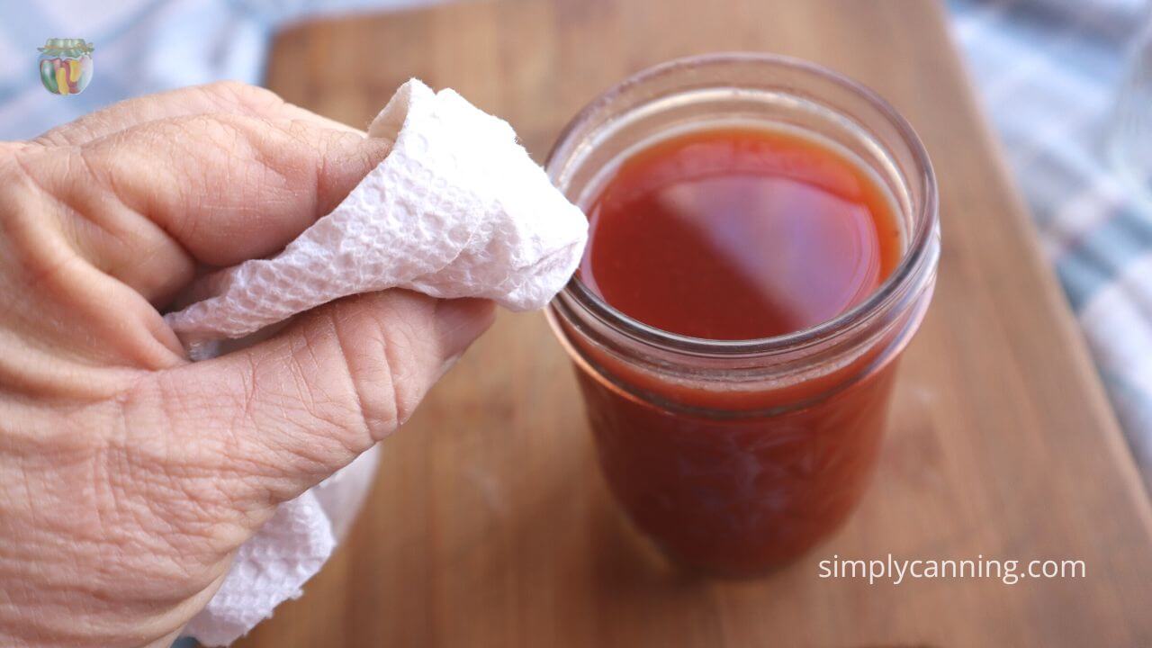 Close up of a jar of tomato juice with the rim being wiped clean with a paper towel. 