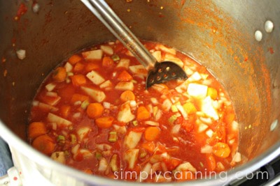 Stirring a big pot of veggie soup that includes potatoes, carrots, and tomatoes.