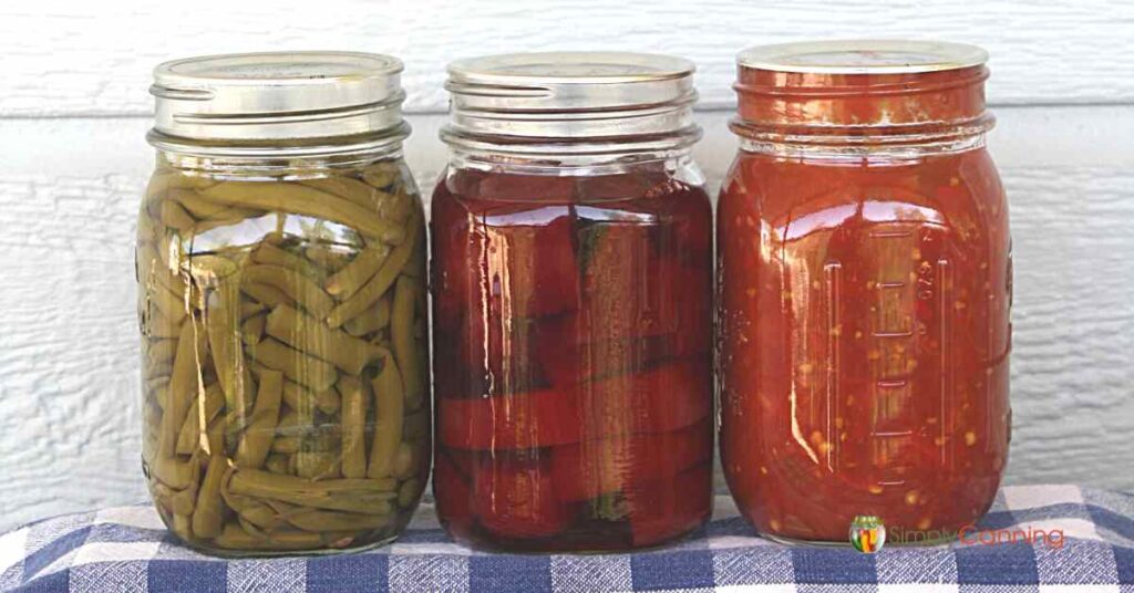 Jars of home canned green beans, beets, and tomatoes sitting on a blue and white checkered napkin.