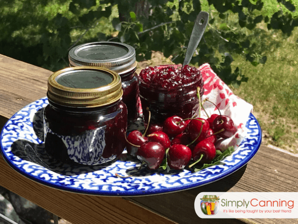 Three jars of homemade cherry jam sitting on a plate with fresh cherries to the side.