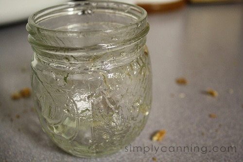 An empty jar with residual dandelion jelly inside and breadcrumbs sprinkled on the countertop around it.