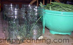 Putting heads of fresh dill into canning jars with a bowl of beans in the background.