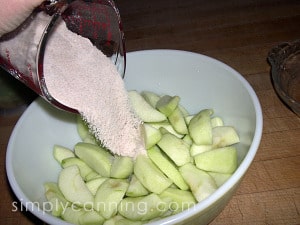 Pouring sugar mixture into a bowl of peeled and sliced apples.