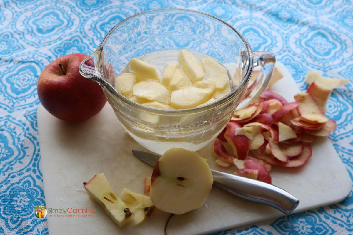 Whole apple, clear mixing bowl with sliced apples, apple peels and a half sliced apple all sitting on a white cutting board.