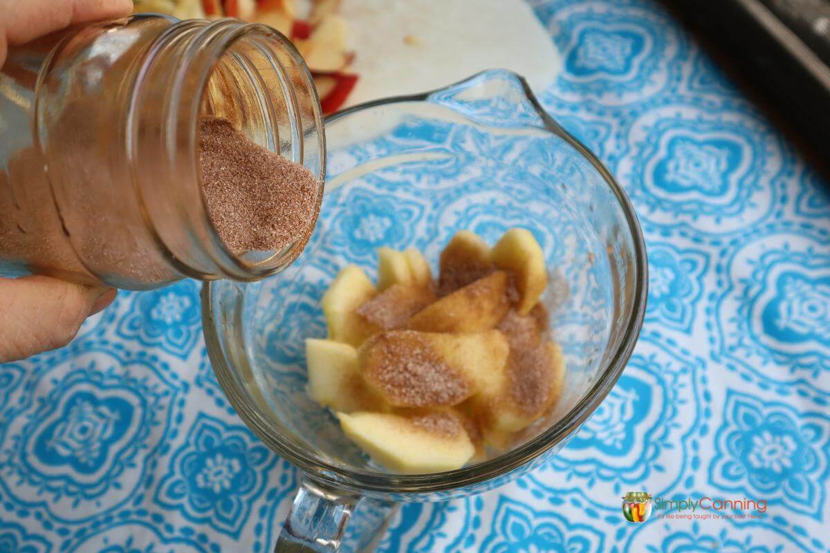 Peeled sliced apples in a clear mixing bowl being mixed with cinnamon sugar.