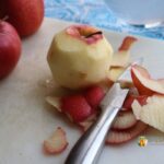 Whole apples sitting on a white cutting board, one of the apples is peeled with the paring knife.