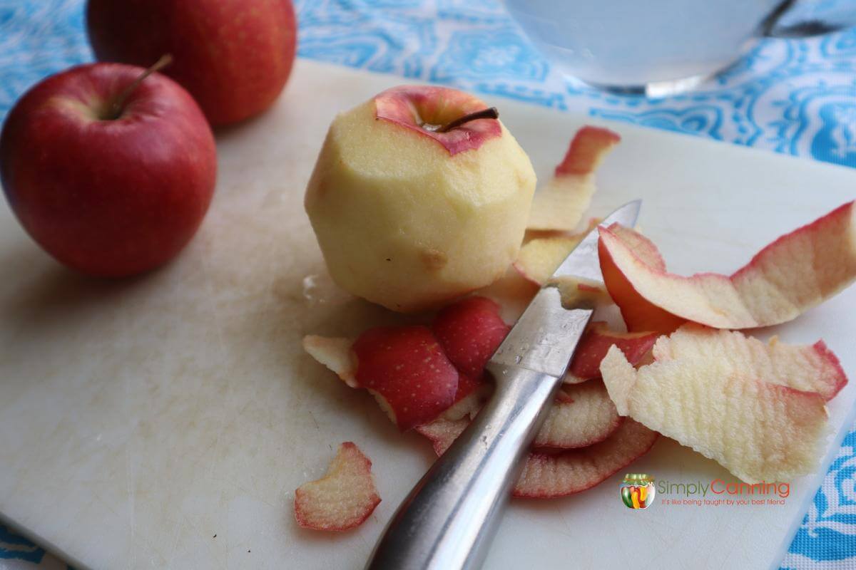 Whole apples sitting on a white cutting board, one of the apples is peeled with the paring knife.