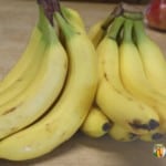 Two bunches of bright yellow bananas with bags of apples in the background.