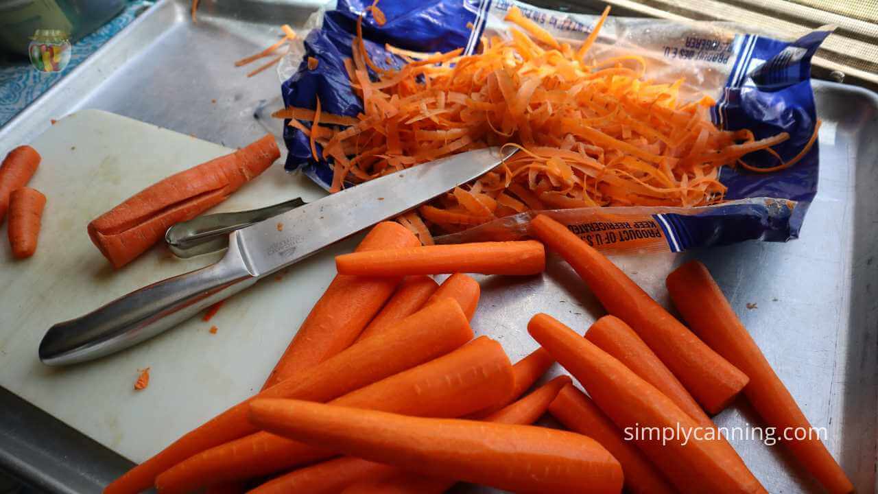 oversized metal tray with small white cutting board, large, knife, vegetable peeler and whole unpeeled carrots piled on the side and freshly peeled carrots piling up. 