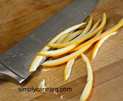 Slicing orange peels into thin strips using a large knife on a wooden cutting surface.