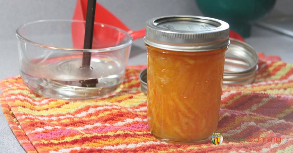 A jar of orange marmalade with various canning supplies around it.