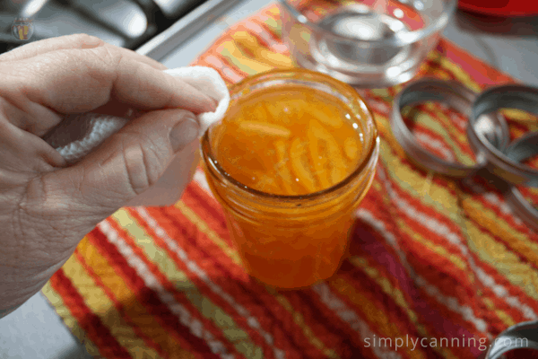 Wiping the rim of a jar of orange marmalade that is sitting on a bright orange striped dishtowel. 