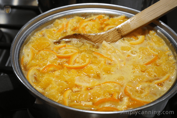 Boiling pot of orange peels and fruit on the stove being stirred with a wooden spoon.