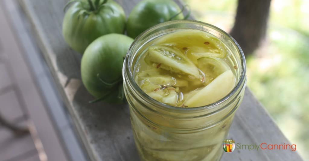 An open jar of pickled green tomatoes with whole green tomatoes in the background.