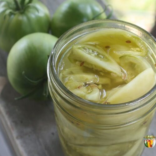 An open jar of pickled green tomatoes with whole green tomatoes in the background.