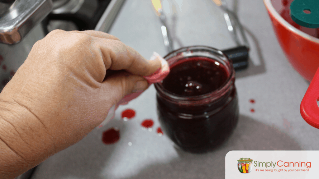 Wiping the rim of a jar of plum jam with a wet paper towel.