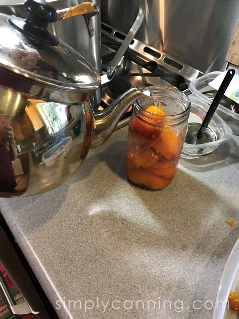 Pouring syrup from the tea kettle into a jar packed with peach halves.