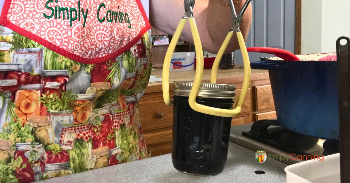 Close up of jar lifter picking up a jar of jam to put in the canner, Sharon in background with her simply canning apron.