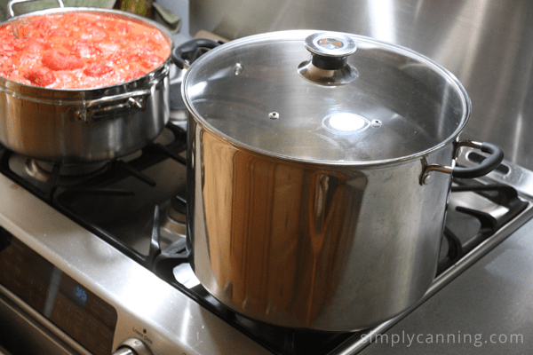 Stainless steel water bath canner sitting on the stove next to a pot of boiling jam.