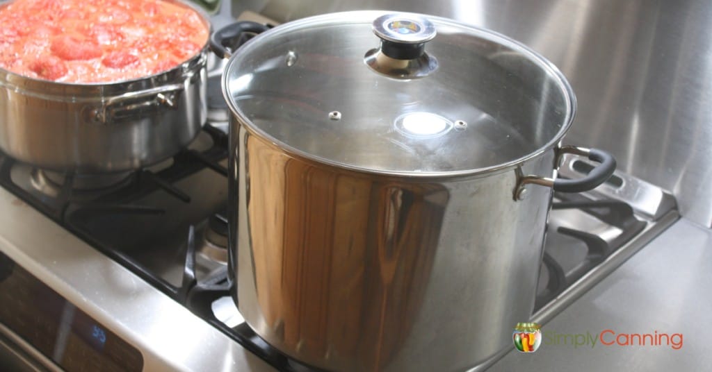 Stainless steel water bath canner sitting on the stove next to a pot of boiling jam.