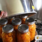 Lifting the lid of a steam canner with jars of peaches in the pot.