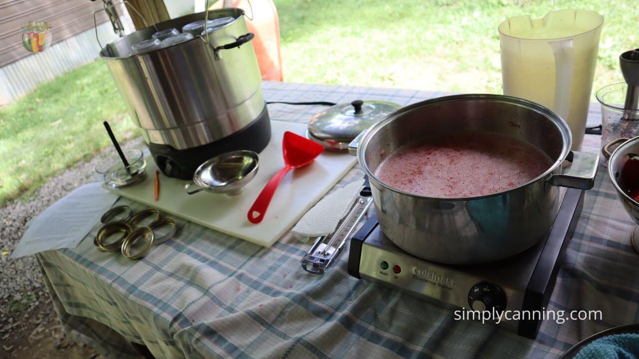 Canning outdoors with ball electric water bath canner and a hot plate heating up some pureed strawberries, various canning supples laid out on the table.   