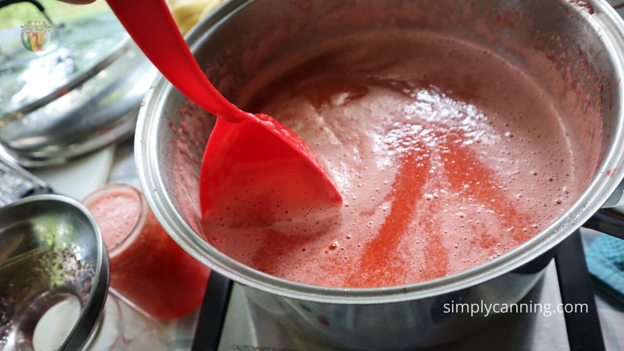 Pot of strawberry concentrate with foam being skimmed by a red 3 sided ladle.