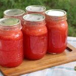 5 jars of homemade strawberry lemonade concentrate sitting on a small cutting board on the picnic table.